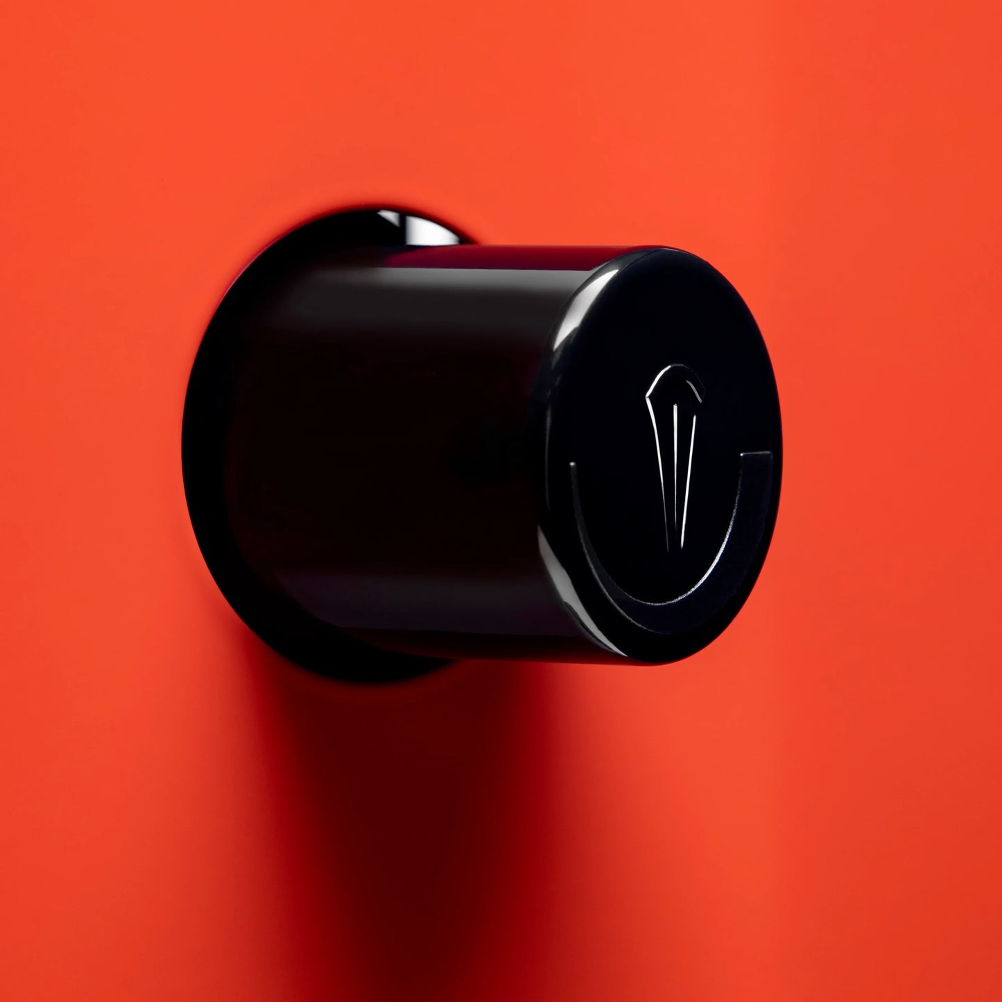 A glossy black cylindrical wall-mounted hook with a stylized silver emblem on the front, set against a solid bright red background.


